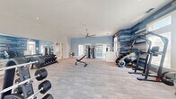 This expansive, single-level fitness center is designed to accommodate a comprehensive range of workout routines across several well-defined zones. The space is bright and airy, benefiting from an abundance of natural light streaming through numerous large windows fitted with blinds, complemented by recessed ceiling lighting and oversized ceiling fans that maintain comfortable airflow throughout.

The main strength training area is generously appointed with a full dumbbell rack, adjustable benches, a multi-station cable machine, a Smith machine, and a dedicated power rack. A wall of floor-to-ceiling mirrors amplifies the sense of space and allows users to monitor their form during lifts. A separate cardio zone features multiple treadmills and stationary bikes, including Peloton-style cycles with integrated screens, all positioned facing windows with outdoor views.

A dedicated functional training room houses kettlebells, medicine balls, a heavy bag, and a pull-up/dip station, along with additional spin bikes. This area features elevated ceilings with a vaulted detail and its own large ceiling fan.

A distinctive geometric, multi-toned blue accent wall serves as a striking architectural focal point in one section of the facility. The flooring transitions between neutral-toned carpet tile and wood-look vinyl, both durable and practical choices for an active environment. Mounted flat-screen televisions are positioned throughout for entertainment during workouts.