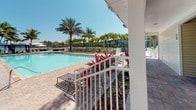 This single-level outdoor amenity space is designed for resort-style leisure and entertaining in a tropical setting. At its centerpiece is a generously sized swimming pool with crystal-clear blue water, surrounded by a broad brick-paver pool deck lined with lounge chairs and accented by colorful tropical plantings, including vivid bromeliads and towering palm trees that frame the open sky above.

Adjacent to the pool, a dedicated covered outdoor kitchen station provides a stainless steel built-in grill housed beneath a clean white gabled roof, making it ideal for outdoor cooking and gatherings. The paver terrace extends to a shaded dining patio furnished with multiple wrought-iron table-and-chair sets, each shaded by large green and striped market umbrellas, creating a comfortable al fresco dining environment.

White-railed walkways and steps connect the various zones of the space, while glass-paneled doors offer direct access to an interior building. The entire area is bathed in abundant natural sunlight under clear blue skies, with the surrounding tropical canopy of mature palms enhancing the relaxed, resort-like ambiance. The cohesive use of brick pavers throughout ties the space together, delivering a polished and inviting outdoor experience.