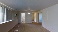 This single-floor space presents an open and functional layout, with the main living area serving as the central hub connecting several adjoining rooms. The living space is finished with hardwood-style flooring that extends throughout, lending a warm, cohesive feel to the interior. A textured ceiling adds subtle depth overhead, while a classic pendant light fixture hangs at the center, providing a focal point and ambient illumination.

Along the left wall, a wall-mounted air conditioning unit offers climate control. A partial counter or bar area is also visible along this wall, hinting at additional utility within the main space. The kitchen, partially visible through an open passageway, features white upper cabinetry, white appliances including a range, and a sink beneath the cabinetry — all contributing to a clean, utilitarian aesthetic.

Toward the back, a hallway leads to additional rooms, with one doorway revealing a carpeted area, suggesting a bedroom or secondary living space. The neutral color palette of soft beige and white walls throughout creates a bright, understated environment that is move-in ready and highly adaptable to various furnishing styles.