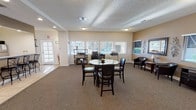 This single-floor communal space offers a bright and functional environment designed for both gathering and productivity. The central focal point is a round dining table surrounded by dark wood chairs with cushioned seats, providing a comfortable area for meetings or casual dining. The textured ceiling and recessed lighting complement the warm, neutral tones of the carpeted flooring throughout the main area.

Along the windowed wall, a dedicated workstation area houses a desk with computer equipment, making the space practical for administrative or business-related tasks. Multiple windows with horizontal blinds line the walls, allowing an abundance of natural light to fill the room while offering views of the surrounding outdoor greenery.

A cozy seating nook in the corner features leather armchairs and a small side table, creating a relaxed lounge area. Decorative wall art adds a touch of character to the otherwise clean and understated aesthetic. A glass-paned entry door on the left side of the space contributes additional light and provides easy access. The overall ambiance is clean, organized, and welcoming, making this a versatile space well-suited for community interaction, work, or relaxation.