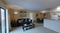 This single-floor space showcases an open-concept design that seamlessly connects the living and dining areas, creating a cohesive and functional layout. The living area is anchored by plush wall-to-wall carpeting in a neutral beige tone, providing a warm and comfortable foundation. A dark upholstered sofa with decorative cushions serves as the focal point, complemented by a glass-and-metal coffee table that adds a contemporary touch. Decorative wall art enhances the neutral beige walls, contributing to the space's modern aesthetic. A stylish floor lamp provides additional ambient lighting alongside the flush-mount ceiling fixture. The textured popcorn ceiling adds subtle visual interest overhead. Adjacent to the living area, the dining space accommodates a sleek table with dark chairs, illuminated by a pendant light fixture. A half-wall or breakfast bar delineates the boundary between the dining area and what appears to be the kitchen beyond. A white interior door leads to additional areas of the unit, maintaining the clean and cohesive design throughout the space.