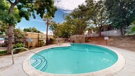Located at 2590 Capitales Dr in Rancho Cordova, California, this single-level outdoor amenity area centers around two distinct swimming pools set within a landscaped community environment. The larger pool features a freeform, curved design with a smooth concrete surround, offering generous deck space ideal for lounging and recreation. Its clear turquoise water is complemented by built-in entry steps and handrail access points. Adjacent to the main pool, a second circular pool provides an additional aquatic option within the same enclosed grounds.

The pool area is framed by mature deciduous trees and a tall palm, providing natural shade and a sense of privacy. A compact utility structure near the poolside serves as a dedicated facility building, and a small patio area with outdoor seating offers a comfortable space for residents to gather. The perimeter is secured by metal fencing, clearly delineating the pool zone from the surrounding property.

The grounds also include an unpaved hillside slope along the rear boundary, lined with chain-link fencing and dotted with established trees. The overall setting reflects a practical, community-oriented outdoor space designed to serve the recreational needs of its residents under the sunny skies of the greater Sacramento area.