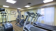 This single-level fitness center provides a functional workout environment equipped with a variety of exercise equipment to support both cardio and strength training. The room features two treadmills positioned near a window dressed with sheer white curtains, allowing natural light to brighten the space. A multi-station cable weight machine occupies the far corner, offering a range of resistance training options, while a stationary bike rounds out the equipment selection for a comprehensive workout experience.

The walls are painted in a warm yellow tone, contributing to an energizing atmosphere, and are accented by framed informational boards and a wall clock. Overhead, recessed panel lighting ensures consistent illumination throughout the room. The floor is covered in a patterned carpet featuring earthy tones of red, green, and gold, adding a decorative element to the practical setting. A rubber mat is placed beneath the weight machine to protect the flooring and provide stability during use. Overall, the space is efficiently organized, making good use of its footprint to offer guests a convenient and accessible fitness facility.