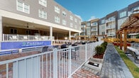 The Parkway amenity space encompasses a resort-inspired outdoor pool area and a comprehensive fitness center, designed to support an active and social lifestyle. The pool deck is finished with brick-paved surfaces and enclosed by a crisp white fence, creating a defined and inviting outdoor retreat. Multiple lounge chairs are arranged poolside, while a covered patio supported by white columns provides shaded seating options. Pergola structures add architectural character to the deck, and the surrounding landscaping introduces greenery to the setting. The pool itself features a bold branded tile detail, reinforcing the community's identity.

Adjacent to the pool, the fitness center is a spacious, light-filled facility with herringbone-patterned flooring and a large ceiling fan overhead. Floor-to-ceiling mirrors line the walls, amplifying the sense of space and aiding in form correction during workouts. The gym is outfitted with a wide range of equipment, including treadmills, ellipticals, cable machines, free weights, a full dumbbell rack, and medicine balls. Built-in wooden shelving offers organized storage for towels and accessories. Large windows and glass sliding doors connect the gym to the outdoors, allowing natural light to flood the interior and offering views of the pool area.