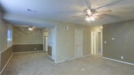 This single-floor space offers a practical and open layout designed for comfortable everyday living. The main living area is carpeted and generously sized, providing ample room for furnishings and movement. A ceiling fan with an integrated light fixture is mounted overhead, contributing to both air circulation and ambient lighting throughout the space.

Neutral beige walls with a textured ceiling create a clean, understated backdrop throughout. A partial dividing wall separates the main living area from an adjoining kitchen space, which is visible in the background and includes a refrigerator. The kitchen area features its own ceiling fan and light combination, maintaining consistent airflow across the open floor plan.

Multiple interior doors lead to additional spaces, including what appears to be a hallway connecting further rooms. A louvered closet door is visible near the kitchen, providing convenient storage access. Floor-level HVAC vents are positioned throughout, ensuring efficient climate control. The overall ambiance is straightforward and functional, with a neutral color palette and practical finishes suited to a variety of living arrangements.