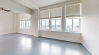Located on the 13th floor at 11 E Forsyth Street in downtown Jacksonville, Florida, this unit offers an efficiently designed open-plan layout that combines the living room and kitchen into one cohesive space. The smooth, polished grey floors extend throughout, lending a modern, clean aesthetic that reflects the abundant natural light pouring in through the large multi-pane windows. These windows, fitted with horizontal blinds, frame impressive panoramic views of the Jacksonville cityscape, making them a true highlight of the space.

The living area is generous and airy for its footprint, with crisp white walls, a ceiling fan, and a pendant light that add functional comfort and character. A curved, half-wall partition elegantly divides the living space from the kitchen, which features warm honey-toned wood cabinetry, a built-in microwave, black refrigerator, and a wide kitchen counter with an integrated sink. The counter extends outward to create a breakfast bar with a wood-accented edge, ideal for casual dining. Overhead fluorescent lighting in the kitchen ensures a bright and practical workspace. The unit's clean, minimalist design and elevated city views make it a distinctive offering in the heart of downtown Jacksonville.