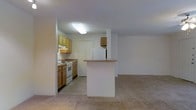 This single-floor space is designed with an open layout that seamlessly connects the kitchen and living area, creating a functional and cohesive environment. The kitchen is equipped with warm honey-toned wood cabinetry offering ample storage, a white electric range, and a full-size refrigerator. A peninsula breakfast bar extends from the kitchen, defining the boundary between the cooking and living zones while providing additional counter space and casual seating potential. Overhead, a flush-mounted fluorescent light fixture illuminates the kitchen area effectively. The countertops feature a neutral laminate surface that complements the cabinetry. The living area is finished with wall-to-wall carpet in a light beige tone, contributing to a comfortable and quiet atmosphere. Walls throughout are painted in a clean, neutral white, enhancing the sense of openness. An additional upper cabinet is positioned on a partial wall near the kitchen, offering supplementary storage. The overall layout is practical and straightforward, making efficient use of the available space on a single level.