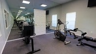 This single-floor fitness center offers a practical and functional workout space designed to support a range of exercise routines. The room is outfitted with multiple pieces of cardio equipment, including a treadmill, an air resistance exercise bike, an elliptical-style stepper, and additional training machines, providing users with diverse options for both low- and high-intensity workouts. Large floor-to-ceiling mirrors line one wall, creating a sense of expanded space and allowing users to monitor their form during exercise. The flooring is covered in durable, low-pile carpet suited for gym use. Bright recessed panel lighting illuminates the space evenly from above, ensuring excellent visibility throughout. A wall-mounted television is positioned for convenient viewing during workouts. Natural light enters through windows fitted with horizontal blinds, offering adjustable light control. The walls are painted in a neutral tone, contributing to a clean and focused atmosphere. A standard door provides access to the room, and the overall layout is open and uncluttered, allowing for comfortable movement between equipment. This fitness center is a well-organized and fully functional space suited for daily cardiovascular and general fitness training.