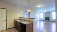 This single-story space showcases an open-concept layout that seamlessly connects the kitchen and living areas, creating a cohesive and functional environment. The kitchen is anchored by a peninsula with granite-look countertops, providing ample prep space and serving as a natural divider between the kitchen and the adjoining living area. Dark cabinetry lines the lower kitchen section, complemented by a double-basin stainless steel sink and a black dishwasher. Above the peninsula, two pendant lights hang at staggered heights, adding a stylish focal point while providing task lighting. A mirrored backsplash element above the sink enhances the sense of depth in the kitchen. The flooring transitions from tile in the kitchen to smooth hardwood-style planks in the living area, adding warmth and visual continuity. The living space benefits from natural light streaming through a window near the entry door, brightening the room considerably. A ceiling fan is visible in the living area, contributing to air circulation and comfort. Neutral wall tones throughout keep the space feeling clean and versatile, while the vaulted ceiling adds a sense of openness and volume to the overall layout.