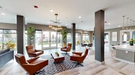Located at 269 S State Highway 360 in Mansfield, Texas, this contemporary community clubhouse offers a thoughtfully designed single-floor layout that balances social gathering spaces with relaxed lounging areas. The entrance foyer features light wood-look flooring, curated artwork on warm taupe walls, and French doors that lead into the heart of the space.

The main lounge area is anchored by rich cognac leather seating arranged over a bold geometric area rug, complemented by a modern sputnik-style chandelier overhead. Structural columns clad in dark charcoal add architectural definition throughout the open floor plan. An adjacent lounge features a dramatic stacked stone accent wall with a wall-mounted flat-screen television, a plush charcoal sofa, and a sculptural marble-top coffee table. Decorative black bamboo stalks add an organic, artistic touch.

The full kitchen is outfitted with dark charcoal cabinetry, warm wood upper shelving, a bright white tile backsplash, and stainless steel appliances. A generous center island with a quartz countertop and gold-accented bar seating serves as a social hub beneath cylindrical glass pendant lights.

Throughout the space, floor-to-ceiling windows and glass doors flood the interior with natural light and provide views of the outdoor courtyard and pergola. Intimate bistro nooks with vibrant yellow accent chairs offer cozy spots for casual conversation or quiet moments.