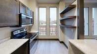 Located at 229 W Third St in downtown Davenport, Iowa, this one-bedroom, one-bathroom unit offers a well-designed layout across a single floor. The kitchen is a standout feature, equipped with dark espresso cabinetry accented by brushed gold hardware, light marble-style countertops, stainless steel appliances including a refrigerator, electric range, and microwave, and a double-basin sink with a gold-tone faucet. Tall, narrow windows frame views of the surrounding historic downtown streetscape, filling the space with natural light. Open floating shelves add a modern, functional touch to the kitchen area. The bedroom is a comfortable, carpeted retreat featuring neutral walls, dark baseboards, bold dark-paneled doors, and a ceiling fan with overhead lighting. The bathroom is finished with light wood-look tile flooring, a dark vanity with gold drawer pulls, a white countertop, a round mirror, and clean neutral walls. A dedicated closet provides additional storage. Throughout the unit, a consistent design palette of dark accents, warm neutrals, and modern fixtures creates a cohesive and contemporary urban living aesthetic.