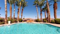 This inviting outdoor amenity space offers a resort-like atmosphere defined by its generous freeform swimming pool, which serves as the centerpiece of the area. The pool's crystal-clear turquoise water is complemented by a decorative rock water feature, adding a naturalistic touch to the landscape. Surrounding the pool, a spacious concrete deck provides ample room for sunbathing, with lounge chairs positioned to take full advantage of the abundant sunshine and open sky views.

Adjacent to the pool, a circular spa offers a more intimate water experience, framed by towering palm trees and classic lantern-style light posts that lend an elegant, resort-inspired character to the setting. Manicured shrubs and greenery are thoughtfully placed throughout, adding color and texture to the landscape.

A covered patio area features a pergola-style shade structure with rustic wooden beams, providing a comfortable retreat from the sun. This lounge space is furnished with wrought iron seating accented by vibrant striped cushions, centered around a coffee table — ideal for relaxing conversation or casual gatherings. The ironwork fencing that encloses the entire area ensures both security and unobstructed views of the palm-lined surroundings and distant desert landscape, reinforcing the open, airy ambiance of this well-appointed outdoor space.