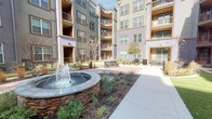 Located at 11701 Mira Lago Blvd in Farmers Branch, Texas, this single-floor residence is part of a well-appointed multi-story community defined by contemporary architecture with warm neutral tones, curved façades, and private balconies. The community's interior courtyard serves as a standout communal feature, centered around a striking stone-base water fountain surrounded by manicured landscaping, ornamental grasses, and paved walkways with seating benches — creating a serene outdoor retreat.

Inside, the unit showcases a clean, modern design with light hardwood-style flooring and a neutral color palette throughout. The kitchen is a functional highlight, equipped with dark gray shaker-style cabinetry, stainless steel appliances including a refrigerator, range, and microwave, and a generous granite-topped island with an undermount sink and pendant lighting — ideal for both cooking and casual dining. The open layout allows natural light to flow through the living and dining areas, which connect seamlessly to the bedroom. The entryway features raised-panel gray doors, including a double closet, lending a polished, cohesive finish. A cozy nook with room for a bistro table adds a versatile touch to the living space.