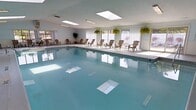 This single-floor indoor pool facility offers a bright and inviting aquatic environment designed for relaxation and recreation. The centerpiece is a generously sized swimming pool with calm, clear water that reflects the surrounding space, giving the room an open and airy feel. The pool deck is lined with wooden Adirondack-style chairs, providing comfortable poolside seating for guests to unwind before or after a swim. Natural light streams abundantly into the space through a row of large windows along one wall, complemented by multiple skylights overhead that further illuminate the interior and reduce the need for artificial lighting. Hanging baskets of lush, trailing ferns and potted tropical plants are thoughtfully placed throughout, adding vibrant greenery that enlivens the clean white walls and ceiling. The overall design is simple and functional, with a well-maintained deck surface surrounding the pool on multiple sides. The white-toned walls and ceiling enhance the brightness of the room, creating a clean and welcoming ambiance. This space is well-suited for both leisure swimming and casual gathering, offering a comfortable, resort-like atmosphere throughout.