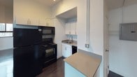 This single-floor unit presents a clean, modern aesthetic anchored by dark hardwood flooring that flows consistently throughout the space. The kitchen is efficiently designed, featuring a full suite of black appliances including a refrigerator, microwave, electric range, and dishwasher, complemented by white cabinetry, a stainless steel sink, and a neutral-toned countertop that extends to a peninsula providing additional prep or dining space. The white walls and recessed layout keep the kitchen feeling bright and organized.

The living areas are open and versatile, with generous wall space and large windows fitted with air conditioning units that frame urban cityscape views, allowing natural light to fill the rooms. Baseboard heating units are present throughout. The bedroom includes a window with a roller shade for privacy and direct access to a built-in closet with shelving, offering practical storage. An additional room provides similar proportions and natural light with city views.

The bathroom is finished with dark tile surrounding a glass-enclosed shower, chrome fixtures and grab bars, a wall-mounted mirror, and a clean white vanity. The overall ambiance of the unit is neutral, polished, and functional, well-suited to urban living.