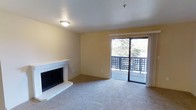 This single-floor residence offers three bedrooms and two bathrooms across a thoughtfully arranged floor plan. The living room serves as the heart of the home, highlighted by a white brick fireplace with a raised hearth that adds warmth and character to the space. Neutral-toned carpet extends throughout the living area, contributing to a soft and comfortable atmosphere. Warm yellow-beige walls complement the light color palette, creating a welcoming ambiance throughout.

A sliding glass door floods the living room with natural light and opens directly onto a private balcony enclosed by a decorative lattice railing, offering views of mature trees and the surrounding outdoor environment. The balcony provides an appealing extension of the living space, ideal for enjoying the outdoors.

The ceiling features a flush-mount light fixture, and a fire sprinkler is also present, reflecting practical safety considerations. The overall design is clean and straightforward, with a neutral finish palette that offers a versatile backdrop suitable for a variety of interior styles. The single-floor layout ensures convenient, accessible living throughout the entire home.