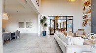 This expansive amenity center is thoughtfully designed across a single floor, offering a diverse collection of spaces suited for socializing, fitness, dining, and focused work. The grand entrance lounge immediately impresses with soaring double-height ceilings, large-format tile flooring, and floor-to-ceiling black-framed glass walls that flood the space with natural light and frame sweeping outdoor views. Plush neutral sofas and curated wall art create a refined yet welcoming atmosphere.

The multiple living areas continue this elevated aesthetic, with one featuring a dramatic floor-to-ceiling slate fireplace surround flanked by contemporary artwork, a mounted flat-screen TV, and sophisticated lounge seating. Another living area boasts a two-story glass wall with panoramic views, anchored by a dark sectional sofa and warm mixed-material furnishings.

The dining and kitchen area is equally impressive, with a large quartz-topped island, dark wood cabinetry with gold hardware, geometric tile backsplash, and stylish pendant lighting. Additional dining spaces offer versatile seating arrangements, from counter-height bar seating to intimate bistro nooks with built-in banquettes.

The two exercise rooms are well-equipped, one housing cardio machines and free weights with panoramic windows overlooking the landscape, and the other dedicated to cycling and functional training with ample natural light. An office rounds out this comprehensive and beautifully appointed facility.