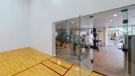 This single-level fitness center offers a comprehensive workout environment designed to accommodate a wide range of exercise needs. The space is anchored by a full-sized racquetball court enclosed with glass walls and a glass-panel door, featuring polished hardwood flooring with regulation court lines. The transparent enclosure allows visibility from the main gym area while maintaining a dedicated, distraction-free playing environment.

The main fitness floor is finished with durable wood-look flooring throughout and is illuminated by recessed ceiling lighting. The cardio zone is equipped with Matrix elliptical trainers, treadmills, and a Peloton cycle, positioned near large sliding glass doors that open to an outdoor patio area, allowing natural light to fill the space and providing views of the surrounding greenery.

The strength training section is generously appointed with a cable functional trainer, a multi-station cable machine, a full rack of dumbbells, adjustable benches, and a heavy punching bag with TRX suspension trainers. Large wall-mounted mirrors expand the perceived depth of the space and allow users to monitor their form. Digital display screens branded with "Perfect Fit" are integrated into the layout. A bold landscape mural adds a dynamic visual element to the strength training area, contributing to an energizing and motivating atmosphere throughout the facility.