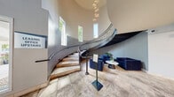 Located at 7201 Walker St in St. Louis Park, MN, this two-floor community space makes a striking first impression with a soaring grand foyer anchored by a sweeping curved staircase. An elegant multi-pendant chandelier cascades from the double-height ceiling, while tall narrow windows flood the entry with natural light. Stone tile flooring and bold navy blue seating arrangements complete the sophisticated welcome area.

The main floor is expansive, offering multiple distinct zones designed for both social gatherings and focused work. A spacious lounge area features a floor-to-ceiling stone fireplace flanked by built-in display niches, a wall-mounted TV, and a mix of plush seating. Adjacent to this, a curated seating nook showcases contemporary artwork and provides a refined transition between spaces. A dedicated work or meeting room, visible through glass doors, features a striking living moss wall accent.

The fitness center is well-appointed with professional-grade strength and cardio equipment, decorative moss panel wall accents, wall-mounted TVs, and mirrored walls that amplify the sense of space. A kitchen area offers stainless steel appliances, white cabinetry, granite countertops, and light hardwood-style flooring with a vibrant blue accent wall.

The upper level, accessible via the iconic curved staircase with ornate wrought-iron railings, provides additional gathering or office space overlooking the grand entry below.
