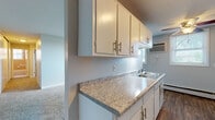 This single-floor unit offers a practical and functional layout across several well-defined rooms. The galley-style kitchen is a standout feature, lined on both sides with white cabinetry equipped with gold-toned hardware and granite-look countertops. A double stainless steel sink, a wall-mounted light fixture, and a white electric range with oven complete the kitchen, which features warm wood-look flooring for a clean, cohesive finish.

The living area is generously sized, with soft neutral carpet, a wide window that allows natural light to fill the room, and a wall-mounted air conditioning unit for climate control. Baseboard heating runs along the perimeter of both the living room and bedrooms, providing efficient warmth throughout.

The bedrooms are carpeted and simply finished with neutral walls, offering a calm and restful atmosphere. One bedroom features a window with views of mature trees outside, adding a touch of natural scenery. Closet space is also present in at least one bedroom.

The bathroom retains a classic character with full wall-to-ceiling pink ceramic tile, a soaking tub with a shower curtain, a window for natural ventilation, and a vanity with mirror. The textured ceilings throughout the unit add an additional layer of detail to the overall interior.
