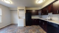 This single-floor residence offers a practical and comfortable layout with a cohesive design throughout. The kitchen is a standout area, featuring rich dark espresso cabinetry with sleek bar-pull hardware, light granite-style countertops, and durable stone-look tile flooring. It is fully equipped with a full suite of black appliances, including a refrigerator, electric range, microwave, and dishwasher, along with a stainless steel sink. A dedicated in-unit laundry nook with a stacked washer and dryer is conveniently integrated into the kitchen area. The living room is generously sized and bathed in natural light from large windows flanking the front entry door. Neutral gray walls and plush wall-to-wall carpeting create a warm and inviting atmosphere. Additional carpeted rooms serve as comfortable bedrooms, each featuring neutral-toned walls and windows providing natural light and outdoor views of surrounding trees. The bathroom is well-appointed with stone-look tile flooring, a white vanity with sink, and a full tub and shower combination with a curved curtain rod. Textured ceilings are present throughout, adding a classic residential character to the space.