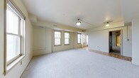 This single-floor space offers a clean, open layout defined by soft neutral walls and a warm, understated aesthetic. The main area is finished with plush wall-to-wall carpeting, creating a comfortable and quiet atmosphere throughout. A row of double-hung windows lines one wall, allowing generous amounts of natural light to fill the interior and lending the space an airy, welcoming feel. The ceiling is smooth and painted white, contributing to the bright, open ambiance, and is complemented by a centrally hung pendant light fixture that adds a subtle industrial touch. Toward the far end of the space, the flooring transitions to hardwood, suggesting a secondary area that could serve a distinct functional purpose. Recessed HVAC vents are integrated into both the ceiling and walls, ensuring consistent climate control throughout. The walls feature clean, flush-panel built-in elements and generous baseboard trim, adding to the tidy, well-maintained character of the space. Overall, the layout is versatile and expansive, with an uncluttered design that makes it highly adaptable for a variety of uses.