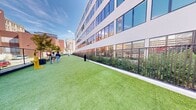 Located at 555 E Main St in downtown Stamford, Connecticut, this outdoor dog run is a thoughtfully designed amenity space set against the backdrop of a modern multi-story residential building. The space features a generously sized, elongated stretch of vibrant artificial turf that provides a clean, low-maintenance surface for pets to roam and play year-round. The run is fully enclosed by a sturdy black metal fence with secure gated entry points, ensuring the safety of all animals within. Raised planting beds line portions of the perimeter, adding greenery and visual appeal with shrubs and ornamental grasses that soften the urban setting. Practical amenities are integrated throughout, including a pet water station and waste disposal stations equipped with bags and receptacles, making the space fully functional for daily use. Young trees are interspersed along the length of the run, offering shade and a natural atmosphere. The surrounding cityscape — including neighboring brick buildings and open sky — gives the space an open, airy feel despite its urban location. The dog run is divided into sections by interior fencing with gated access, allowing for flexible use. Overall, this space offers a well-maintained, accessible, and inviting outdoor environment for pets and their owners in the heart of downtown Stamford.
