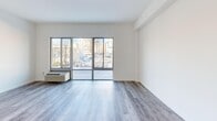 Located at 100 Evergreen Place within The Althea in East Orange, NJ, this single-floor unit offers a thoughtfully designed layout that maximizes its compact footprint. The combined kitchen and living area forms the heart of the space, where large floor-to-ceiling windows flood the room with natural light and frame views of the surrounding East Orange cityscape. Light gray hardwood-style flooring extends throughout the living area, lending a clean, contemporary feel to the space.

The kitchen is efficiently appointed with dark espresso cabinetry, granite countertops, stainless steel appliances including a refrigerator, gas range, and built-in microwave, and track lighting overhead. Neutral-toned tile flooring grounds the kitchen and contrasts neatly with the wood flooring in the adjacent living area.

A dedicated bedroom area features the same hardwood-style flooring and crisp white walls, offering a bright, neutral canvas. The bathroom is finished in a clean, modern style with marble-look tile flooring, a white vanity with an integrated sink, a framed mirror, and white wall finishes throughout. Multiple hallways connect the rooms, contributing to an organized, functional flow throughout the unit.