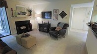 This single-floor living space offers a comfortable and well-appointed layout centered around a classic fireplace, which serves as a natural focal point along one wall. The living area is finished with plush wall-to-wall carpeting in a neutral tone, contributing to the warm and inviting atmosphere throughout. A wall-mounted flat-screen television is positioned above a dark wood media console with glass-front storage, providing both function and style. Decorative framed artwork and an ornate diamond-shaped wall accent add visual interest to the neutral-painted walls. A richly upholstered leather armchair with nailhead trim and carved wooden legs offers an elegant seating option, complemented by a cushioned ottoman that doubles as a coffee surface. A tall floor lamp provides warm ambient lighting, while a shelf above the fireplace displays decorative vases and greenery, adding a touch of natural detail. An adjacent hallway visible in the background suggests additional rooms beyond, offering a sense of connected, functional living. Overall, the space balances classic design elements with everyday comfort.