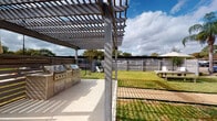 Located at 2121 Tannehill Drive in Houston, TX, this outdoor amenity space is thoughtfully designed to offer residents a functional and inviting environment for relaxation and socializing. The centerpiece of the space is a pergola-covered outdoor kitchen area, equipped with a stainless steel built-in grill and ample counter space, ideal for outdoor cooking and entertaining. The slatted pergola overhead provides partial shade while allowing natural light and open sky views to filter through.

Adjacent to the grill station, a generous open lawn extends across the grounds, offering a versatile green space for leisure activities. A gravel-lined pathway winds through the yard, connecting various seating areas that feature Adirondack chairs and picnic-style benches, some shaded by a large market umbrella. The lawn is enclosed by horizontal-slat privacy fencing, creating a well-defined and secure perimeter.

A dedicated dog run area is also present, enclosed with chain-link fencing and featuring young trees for light landscaping. Covered carport parking is visible at the rear of the property, adding practical convenience. Mature trees, including a striking palm, frame the space and contribute to its approachable, community-oriented atmosphere under the open Houston sky.