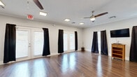 Located at 1407 Autumn Way in Dickson, Tennessee, this single-floor commercial space offers a well-designed, open-concept layout ideal for a variety of uses including classes, gatherings, or events. The main open room is the centerpiece of the space, showcasing rich hardwood flooring that extends throughout and reflects the natural light that pours in through multiple sets of French doors and windows dressed with black curtains for privacy and light control. Two ceiling fans provide comfortable airflow, while recessed flush-mount lighting ensures consistent illumination. Crown molding and clean white walls contribute to a polished, neutral aesthetic. An emergency exit and safety signage reflect the space's commercial-grade design. Adjacent to the main room is a compact but fully functional kitchenette, outfitted with white shaker-style cabinetry, a white countertop island, stainless steel appliances including a refrigerator, microwave, and dishwasher, and a utility sink. Crown molding carries through into this area as well, maintaining a cohesive look. The space also accommodates stacking chairs and folding tables, making it highly adaptable for varied configurations and event setups.