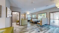 This single-floor communal space is thoughtfully designed to serve as a functional and inviting hub for residents or guests. The interior features wood-look luxury vinyl plank flooring throughout, lending warmth and continuity across all areas.

The main lounge area is generously proportioned, furnished with a mix of upholstered booth-style seating and accent chairs in earthy tones of green, gold, and gray. Large, framed windows line the exterior walls, flooding the space with natural light and offering pleasant views of the surrounding landscaped grounds, including mature trees and tropical foliage. Pendant globe light fixtures provide soft, ambient illumination overhead.

Adjacent to the lounge is a dedicated business center, equipped with desktop computers at a built-in countertop workstation, illuminated by elegant chandelier-style accent lighting. A glass door connects directly to the outdoor courtyard, further enhancing the sense of openness.

A private meeting room offers a more intimate setting, outfitted with a round pedestal table, comfortable seating, and a wall-mounted flat-screen television — ideal for small gatherings or presentations. A window provides natural light to this compact yet functional room.

Decorative touches, including a mosaic wall art piece and thoughtful accent lighting, add personality and polish throughout, creating an atmosphere that balances professionalism with comfort.