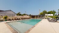 This outdoor pool area is designed for both leisurely relaxation and active recreation, centered around a generously sized rectangular swimming pool with crystal-clear turquoise water. The pool deck is finished in light-toned concrete pavers, providing ample space for lounging and movement around the perimeter. Along one side of the deck, a row of cushioned chaise lounges paired with pink market umbrellas offers a comfortable sunbathing area, while potted palm trees add a tropical touch throughout the space.

A standout feature of the area is a covered pergola structure situated at one end of the pool. Built with timber-frame construction and a pitched roof, this shaded pavilion houses a seating arrangement that includes lounge sofas and dining chairs, making it a versatile retreat for both casual gatherings and outdoor dining.

The pool area is enclosed by a combination of wrought iron fencing and substantial stacked stone retaining walls, which provide privacy and a sense of structure to the space. An adjacent building with a gabled roof serves as a facility structure, likely housing restrooms or pool support amenities. The overall ambiance is open and sun-drenched, with clear sightlines to the surrounding neighborhood and mature trees visible beyond the perimeter.