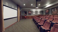 This private home theater is designed to replicate the experience of a classic cinema with thoughtful attention to detail. The room is finished with gray-painted walls accented by rich wood wainscoting that lines the lower half of the perimeter, adding warmth and a traditional, upscale character. Recessed ceiling lighting and decorative wall sconces provide ambient illumination, while crown molding adds a refined architectural touch.

The seating area features multiple rows of plush, red upholstered theater-style chairs equipped with armrests and cup holders, arranged to provide clear sightlines to the screen. A ceiling-mounted projector delivers images onto a large projection screen framed by floor-length tan curtains, creating a true cinematic atmosphere. Framed movie posters are displayed along the side walls, reinforcing the theater aesthetic.

Patterned carpeting covers the floor throughout the space, contributing to both acoustics and visual appeal. At the rear of the room, a built-in bookcase and storage cabinetry provide functional display and organizational space. A classic red popcorn cart near the entrance adds a charming, authentic movie-going touch. The overall design balances functionality with a polished, cinema-inspired ambiance.