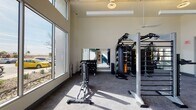 This fitness center in Mesa, Arizona, is a well-appointed, single-floor facility designed to support a wide range of workout styles. The space is characterized by soaring ceilings with exposed structural elements and expansive floor-to-ceiling windows along multiple walls, flooding the interior with abundant natural light and offering views of the surrounding landscaping and courtyard.

The layout is open and spacious, with durable, light-toned flooring throughout. Cardio equipment—including treadmills, upright cycles, and a stair climber—is positioned along the large glazed walls to take advantage of the outdoor views. Strength training is well-represented by a multi-functional rig system equipped with a punching bag, TRX suspension trainers, and cable attachments, along with a dedicated dumbbell rack visible in mirrored wall sections that enhance the sense of depth. Functional fitness accessories, including a heavy tire and plyo boxes, add versatility.

A standout design feature is the bold, large-scale graphic mural in navy, forest green, and white, which adds a dynamic, energetic aesthetic to the space. Ceiling fans and linear pendant lighting complement the natural light. The facility connects to adjacent interior spaces through a glazed door, suggesting access to additional amenities nearby.