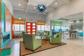 This single-story space in Columbus, Georgia, makes an immediate impression with its bold, energetic color palette and thoughtfully designed layout. The main lounge area serves as the heart of the space, featuring soaring beamed ceilings and recessed lighting that illuminate a seating arrangement of plush lime-green wingback chairs accented with vibrant teal and yellow throw pillows. A gold-framed glass coffee table anchors the grouping atop a woven area rug, while striking red French doors at the entrance flood the room with natural light. Abstract yellow artwork displayed in clean white frames adds artistic flair to the bright white walls. Adjacent to the lounge, a dining nook with teal upholstered booth seating and compact tables offers an intimate gathering spot. A sliding orange barn door leads to a private office area, adding a rustic yet playful architectural detail. The utility kitchen features an extended run of vivid turquoise cabinetry topped with bright white countertops and a farmhouse-style apron sink, all set against a bold geometric patterned tile backsplash. Throughout, the light wood-look flooring and white walls serve as a neutral canvas that allows the vibrant furnishings and design elements to take center stage, creating an inviting and lively atmosphere.
