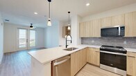 Located in Farmers Branch, TX, this thoughtfully designed one-bedroom, two-bathroom residence offers a clean, modern aesthetic across a well-utilized single floor. Light wood-tone flooring flows consistently throughout, complementing the bright white walls and creating a warm, cohesive atmosphere.

The open-concept kitchen and living area serves as the heart of the home. The kitchen is outfitted with light wood cabinetry, a gray subway tile backsplash, stainless steel appliances including a refrigerator, range, and microwave, and a spacious island with a white countertop and pendant lighting above. A built-in desk nook adds functional workspace to the layout.

The generously sized primary bedroom features a ceiling fan, a large window with outdoor views, and French doors that open to a private balcony, filling the room with natural light.

Both bathrooms reflect the same modern sensibility. The primary bath includes a floating wood vanity with a white countertop, black-framed mirror, black hardware accents, and large-format beige marble-style tile surrounding a soaking tub with built-in corner shelving. Light wood flooring ties the space together seamlessly.

A dedicated closet provides practical storage, rounding out this well-appointed and stylish residence.