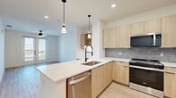 Located in Farmers Branch, TX, this thoughtfully designed one-bedroom, two-bathroom residence offers a clean, modern aesthetic across a well-utilized single floor. Light wood-tone flooring flows consistently throughout, complementing the bright white walls and creating a warm, cohesive atmosphere.

The open-concept kitchen and living area serves as the heart of the home. The kitchen is outfitted with light wood cabinetry, a gray subway tile backsplash, stainless steel appliances including a refrigerator, range, and microwave, and a spacious island with a white countertop and pendant lighting above. A built-in desk nook adds functional workspace to the layout.

The generously sized primary bedroom features a ceiling fan, a large window with outdoor views, and French doors that open to a private balcony, filling the room with natural light.

Both bathrooms reflect the same modern sensibility. The primary bath includes a floating wood vanity with a white countertop, black-framed mirror, black hardware accents, and large-format beige marble-style tile surrounding a soaking tub with built-in corner shelving. Light wood flooring ties the space together seamlessly.

A dedicated closet provides practical storage, rounding out this well-appointed and stylish residence.