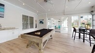 Located at 325 Marymeade Dr in Summerville, SC, this single-floor amenity space is thoughtfully designed for recreation and socializing. The interior is finished with crisp white shiplap walls, light hardwood-style flooring, and recessed lighting throughout, creating a bright and welcoming atmosphere.

The game room serves as a central gathering area, anchored by a rustic wood-framed billiard table with a dark felt surface. The open layout provides ample room for play and movement, while large glass-paned doors connect seamlessly to adjacent areas, enhancing the flow between spaces.

A well-appointed wet bar area features granite countertops, white cabinetry, a stainless steel sink, a beverage cooler, and a dedicated ice machine — all positioned beneath a wall-mounted television, making it an ideal entertainment hub.

The lounge area is defined by full-height glass panel garage doors that open completely to the outdoors, offering direct views of the community pool and surrounding greenery. A high-top dining table with seating is positioned to take full advantage of the indoor-outdoor connection, allowing natural light to flood the space and providing easy access to the pool deck.

Together, these areas form a versatile, resort-style community space well-suited for gatherings, leisure, and everyday amenity use.