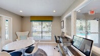 Located at 569 Little River Loop in Altamonte Springs, Florida, this single-floor community clubhouse is designed with a clean, contemporary aesthetic that balances functionality and comfort. The open-plan layout is anchored by a spacious lounge area featuring crisp white sofas accented with mustard yellow and teal throw pillows, a glass-top coffee table, and a statement sputnik chandelier overhead. Light-toned large-format tile flooring runs throughout, enhancing the bright and airy atmosphere. Recessed lighting complements abundant natural light streaming in through multiple windows and glass doors that open directly to the outdoor pool area, seamlessly connecting indoor and outdoor living. A built-in refreshment bar with white cabinetry, a beverage cooler, and countertop appliances provides convenient amenities for residents. A dedicated business center features multiple computer workstations with ergonomic seating, a printer, and large mirrors that visually expand the space. An adjacent seating nook includes a round marble-top table with modern white chairs, ideal for casual meetings or dining. A wall-mounted flat-screen TV and a stylish egg chair create a comfortable media corner. Framed artwork and decorative accents throughout add personality, making this clubhouse a well-appointed and inviting community hub.