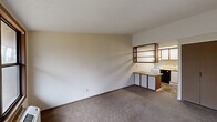 Located at 3052 Orchard Glen Dr in Dayton, Ohio, this single-floor unit offers a practical and efficient layout across three functional spaces. The living area features neutral-toned carpet flooring, warm beige walls, and a textured ceiling, creating a simple and clean environment. A large window with wood-trimmed framing allows natural light to filter in, accompanied by a wall-mounted HVAC unit for climate control and a door providing direct outdoor access.

The kitchen is positioned adjacent to the living area and is accessible via a transition to tile flooring. It is equipped with white upper cabinetry accented by dark wood trim, a compact electric range, a countertop with a stainless steel sink, and a mini refrigerator below the counter. Open shelving above the kitchen peninsula offers additional storage and visual separation between spaces. Overhead fluorescent lighting illuminates the kitchen area effectively.

The bathroom includes a single vanity with a built-in sink, a toilet, towel bars, and a bathtub, all finished with tile flooring that complements the kitchen. A dedicated laundry area rounds out the unit's functionality. Overall, the space presents a straightforward and utilitarian design, well-suited for single-occupant living in a convenient Dayton location.