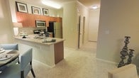 This single-floor residence showcases an open-concept layout that seamlessly connects the kitchen and living areas. The kitchen is equipped with stainless steel appliances, including a refrigerator, microwave, and range, complemented by warm medium-toned wood cabinetry extending to the upper walls. A peninsula with a granite-style countertop and integrated sink serves as a functional divider between the kitchen and the adjoining carpeted living space, offering additional prep and casual dining space. Crown molding adds a refined architectural detail along the ceiling, enhancing the overall finish of the space. The neutral beige wall tones and light-colored carpet create a cohesive, understated palette throughout. A hallway extends from the main living area, leading to additional rooms and a closet with louvered doors, suggesting practical storage solutions. The entryway door is visible at the end of the corridor, reinforcing the single-floor, straightforward layout. Flush-mount ceiling lighting provides consistent illumination across the kitchen and hallway, contributing to the bright and functional character of the space.