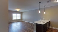 This single-floor space showcases an open-concept layout that seamlessly connects the kitchen and living area, creating a cohesive and functional environment. The kitchen features a dark-cabineted island with a light-colored countertop, an undermount sink with a modern arc faucet, and two sleek pendant lights hanging overhead, adding a contemporary touch to the space. The living area is defined by warm hardwood-style flooring that runs throughout, complementing the neutral gray wall tones and crisp white baseboards. A double window along the far wall invites natural light into the living space, enhancing its open and airy feel. A flush-mount ceiling fixture provides additional ambient lighting in the living area. The overall design balances modern aesthetics with practical functionality, offering a clean, neutral palette that allows for versatile interior styling. The unobstructed sightlines between the kitchen and living area make the space feel expansive, while the cohesive flooring and color scheme tie the two areas together harmoniously.