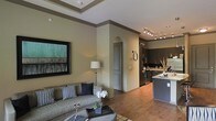 This single-floor space showcases an open-concept layout that seamlessly connects the living area and kitchen, creating a cohesive and functional environment. The living area is anchored by warm hardwood flooring, which extends throughout the space, complementing the earthy, neutral wall tones. A tray ceiling with crown molding adds architectural interest and a sense of elevated elegance to the room. The living area is well-appointed, featuring ample space for comfortable seating and accented by a large framed artwork on the wall, contributing to a polished aesthetic. A floor lamp provides warm ambient lighting, enhancing the inviting atmosphere. An arched doorway serves as a stylish architectural transition between the living space and the adjoining areas. The kitchen is defined by rich, dark cabinetry that provides a striking contrast against lighter countertops, including a functional island. Track lighting illuminates the kitchen workspace efficiently, while modern appliances complete the well-equipped culinary space. The overall ambiance of this space is sophisticated yet comfortable, with thoughtful design details and a harmonious blend of textures and tones throughout.