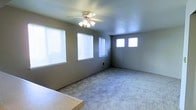 This single-floor space features a generously sized main living area finished with wall-to-wall light gray carpeting, providing a soft and comfortable underfoot experience. The room is painted in neutral gray tones, creating a calm and versatile backdrop suitable for a variety of interior styles.

Natural light is a notable feature of this space, with multiple windows fitted with white horizontal blinds lining the walls. These windows allow abundant daylight to fill the room from multiple angles, enhancing the open and airy feel of the interior.

A centrally mounted ceiling fan with an integrated light fixture serves as both a functional and practical element, providing air circulation and ambient lighting throughout the space.

A wall-mounted heating unit is visible, ensuring year-round climate comfort. The room also includes a countertop or partition element visible at the foreground, suggesting proximity to a kitchen or breakfast bar area.

The overall layout is open and unobstructed, offering flexibility for furniture arrangement. The combination of neutral finishes, ample natural light, and practical fixtures makes this a straightforward and functional living environment.