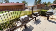 This single-story outdoor amenity space is designed to serve as a communal gathering area, offering residents a well-equipped environment for outdoor dining and recreation. The space is divided into two primary functional zones, each serving a distinct purpose.

The covered patio area features a generous concrete slab shaded by a solid stucco-finished roof supported by open-sided posts, allowing natural light and breezes to flow through freely. Beneath the cover, multiple round glass-top tables with blue mesh patio chairs provide comfortable seating for groups. Two doors with bright blue accents are visible along the back wall, likely providing access to restroom or utility facilities. Overhead fluorescent lighting ensures usability beyond daylight hours.

Adjacent to the patio, a dedicated grilling station houses two full-sized gas BBQ grills with side shelving, positioned along the decorative wrought iron perimeter fence. The fencing encloses the entire area while maintaining open sightlines to the surrounding well-maintained lawn, mature trees, and nearby buildings with characteristic red tile roofs and earth-toned stucco exteriors. The overall ambiance is practical and community-oriented, ideal for outdoor social gatherings.