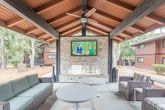 Located at Glenwood Circle in Monterey, California, this single-level outdoor lounge and common area offers a well-appointed gathering space designed for comfort and community. The centerpiece of the space is a covered lounge pavilion featuring a dramatic vaulted wood-beam ceiling with dual ceiling fans, a floor-to-ceiling stacked stone fireplace, and a mounted flat-screen television — creating an inviting atmosphere for year-round outdoor enjoyment. Comfortable wicker seating with teal cushions, along with white accent tables, fills the covered area, providing ample space for relaxation and socializing. The pavilion opens directly onto a spacious concrete patio furnished with additional seating, umbrella tables, and lounge chairs, extending the usable outdoor area. Mature trees, including striking specimens draped with Spanish moss, frame the grounds and provide natural shade. Mulched landscaping borders the pathways that wind through the property, connecting the lounge area to other amenities including a laundry facility and the recently opened Monterey Pines Gym. The warm brown wood-panel architecture of the surrounding buildings creates a cohesive, naturalistic aesthetic throughout the community grounds.