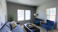 This single-story space features a well-appointed living area designed with a clean, contemporary aesthetic. The room is finished with neutral gray walls that provide a versatile backdrop, complemented by wall-to-wall carpeting that adds warmth underfoot. Two generously sized windows along the exterior wall allow an abundance of natural light to fill the space while offering views of the surrounding greenery outdoors.

The living area is thoughtfully arranged with a gray sofa and a pair of deep navy blue wingback accent chairs, contributing a bold pop of color against the muted palette. A dark-toned rectangular coffee table serves as a central anchor, styled with decorative objects atop a serving tray. A slender floor lamp positioned in the corner provides additional ambient lighting, while a small side table beside the accent chairs offers functional surface space.

The room's overall ambiance is modern yet approachable, balancing practicality with refined style. The consistent use of contrasting dark furniture against lighter walls and flooring creates a visually cohesive environment. The single-floor layout contributes to an accessible, open feel throughout the space.