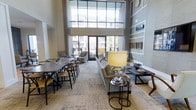 This single-floor space makes a strong architectural impression with its dramatic double-height ceilings and expansive floor-to-ceiling windows framed in dark steel, flooding the interior with natural light and offering views of the surrounding outdoor environment. The open layout seamlessly integrates a lounge area and a dining zone, creating a versatile, multi-functional environment. The lounge section is anchored by a neutral-toned sofa paired with a sleek coffee table and a statement table lamp featuring a warm yellow shade on a metallic base, lending the space a refined yet approachable character. A built-in shelving unit lines one wall, providing both display and storage opportunities. The dining area accommodates a long, dark wooden table surrounded by modern wishbone-style chairs, ideal for group gatherings. Soft, patterned carpeting unifies the space underfoot, while the light grey walls and carefully curated décor contribute to a clean, contemporary aesthetic. Glass doors open directly to the outdoors, enhancing the sense of connectivity between interior and exterior spaces. Overall, the space balances modern design with a welcoming, communal ambiance.