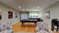 This single-floor recreational space is thoughtfully designed to serve as a versatile entertainment and social hub. The open layout is anchored by a full-size billiards table positioned at the center, set atop warm hardwood flooring that extends throughout the entire space. Recessed lighting illuminates the room evenly, while a set of stylish pendant lights hang directly above the pool table area, adding a focused and inviting ambiance.

Along one wall, a wet bar with granite countertops and dark cabinetry provides a convenient refreshment station, complemented by framed artwork displayed above it. A cue rack nearby keeps the space organized and ready for play.

The lounge area, situated toward the windowed end of the room, is furnished with rich dark leather sofas accented with decorative pillows, patterned accent chairs, and a glass-top coffee table, all arranged atop a neutral area rug. A large window dressed with striped drapes allows natural light to filter in, brightening the seating area. A wall-mounted flat-screen television is positioned for comfortable viewing from the lounge seating, alongside a compact dining table and chairs for more casual gatherings.

Decorative movie posters and framed prints are featured throughout, adding character and a lively, curated aesthetic to the overall space.